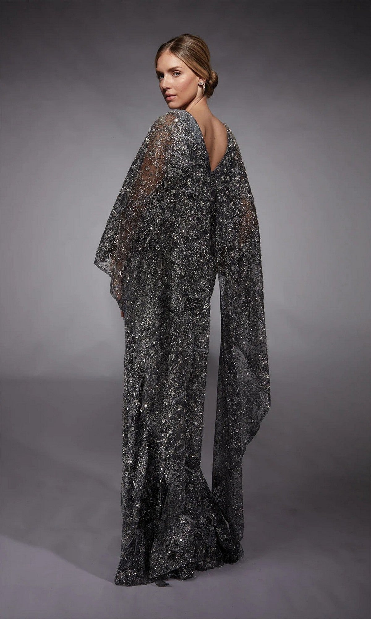 A woman faces away, wearing the Alyce 27752: a floor-length silver glitter tulle formal dress with sheer flowing sleeves, set against a gray backdrop.